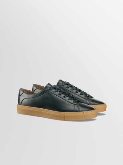 A men's low-top Italian leather sneaker in dark green with a tan gum sole, shown in a side profile view against a light gray background.