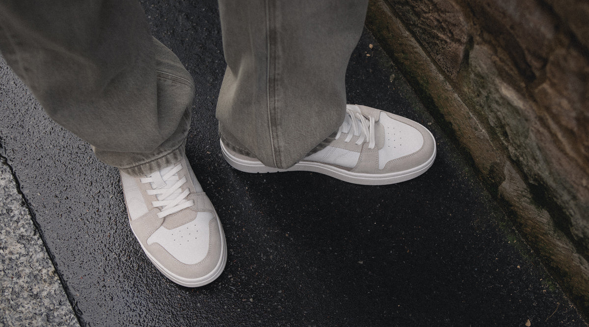 A close-up of a person's feet wearing men's light gray European leather and suede white low-top sneakers, standing on wet dark asphalt next to a stone curb.